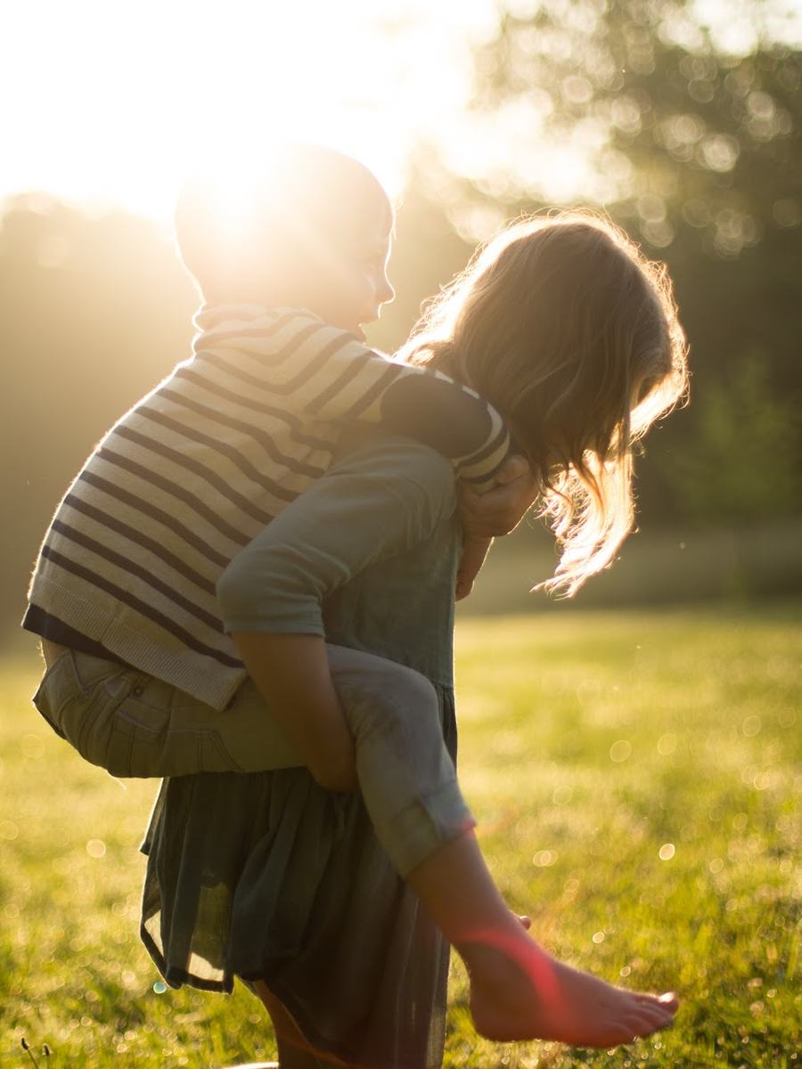 A parent and child playing in an open, sunlit field of grass.